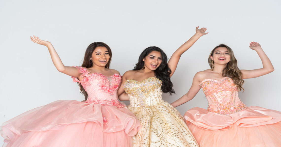 Three teen girls stand together wearing elaborate quinceañera ball gowns with full skirts in a bright outdoor setting.