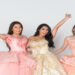 Three teen girls stand together wearing elaborate quinceañera ball gowns with full skirts in a bright outdoor setting.