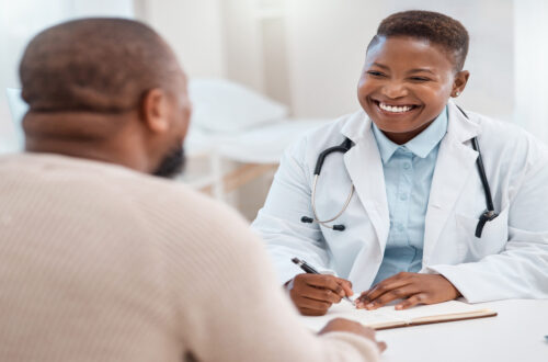 An urgent care doctor wearing a white coat and a stethoscope smiles at a patient. She is ready to write notes on a clipboard.