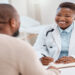 An urgent care doctor wearing a white coat and a stethoscope smiles at a patient. She is ready to write notes on a clipboard.