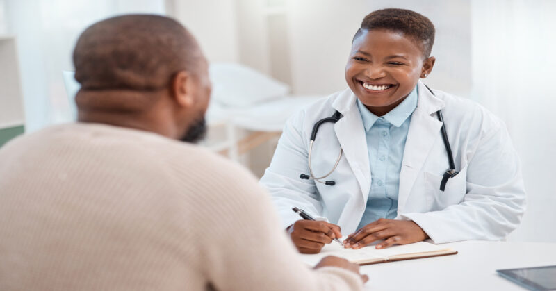 An urgent care doctor wearing a white coat and a stethoscope smiles at a patient. She is ready to write notes on a clipboard.