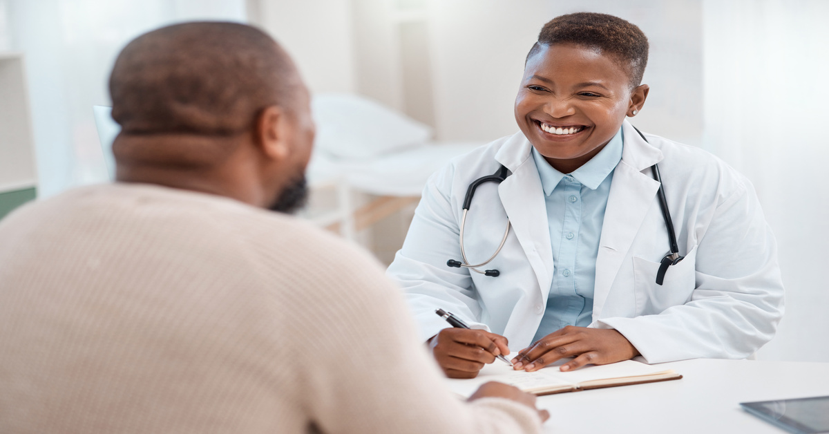 A doctor wearing a white coat and a stethoscope smiles at a patient. She is ready to write notes on a clipboard.