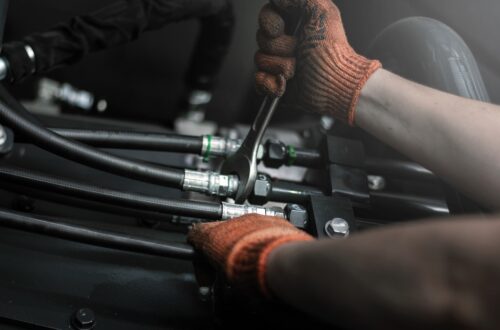 A close-up of someone wearing dirty orange work gloves while tightening an industrial hydraulic fitting.