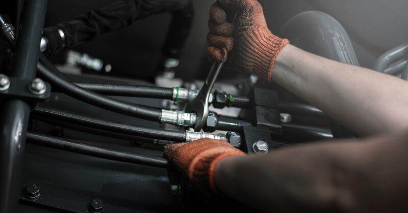 A close-up of someone wearing dirty orange work gloves while tightening an industrial hydraulic fitting.