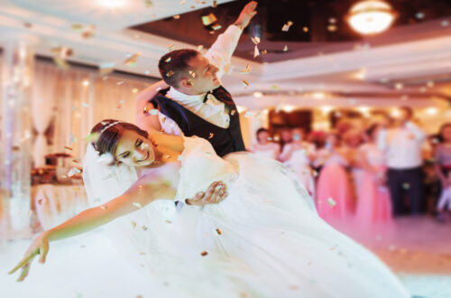 A bride and groom smile as he dips her on the dance floor. Confetti falls from the ceiling, and guests are in the background.