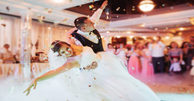 A bride and groom smile as he dips her on the dance floor. Confetti falls from the ceiling, and guests are in the background.