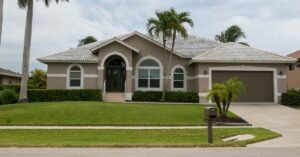 A street view of a single-story, tan-stucco home with palm trees in the yard, a wooden front door, and a brown two-car garage. model home