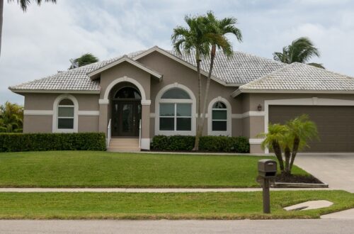 A street view of a single-story, tan-stucco home with palm trees in the yard, a wooden front door, and a brown two-car garage.