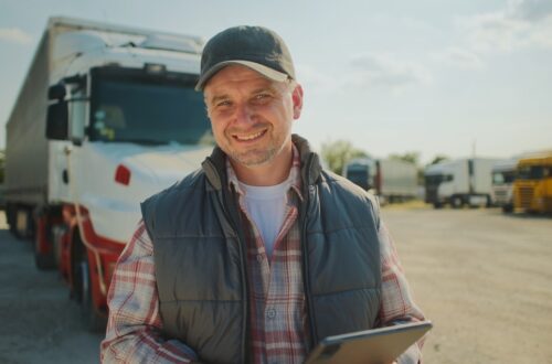 A trucker smiles while standing in front of his parked semitrailer. He holds a tablet and wears a black cap and puffy vest.