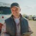 A trucker smiles while standing in front of his parked semitrailer. He holds a tablet and wears a black cap and puffy vest.