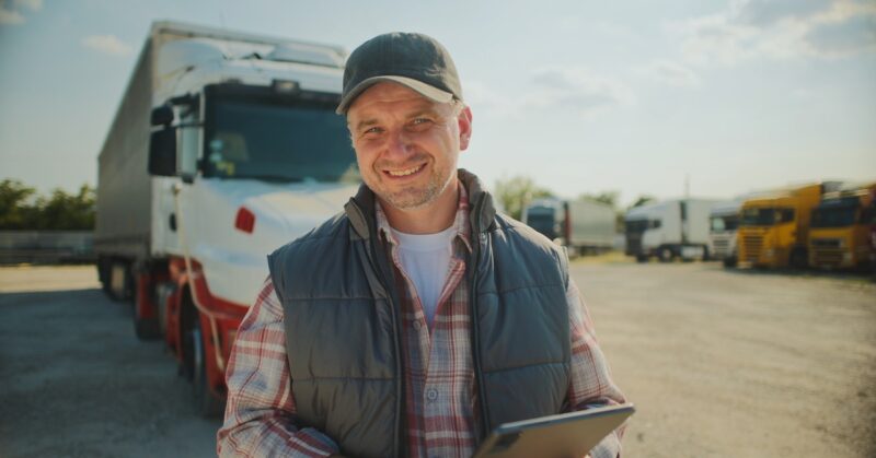 A trucker smiles while standing in front of his parked semitrailer. He holds a tablet and wears a black cap and puffy vest.
