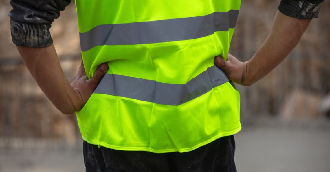 The view of someone from behind, with their hands on their hips, wearing a yellow high-visibility uniform during the day.