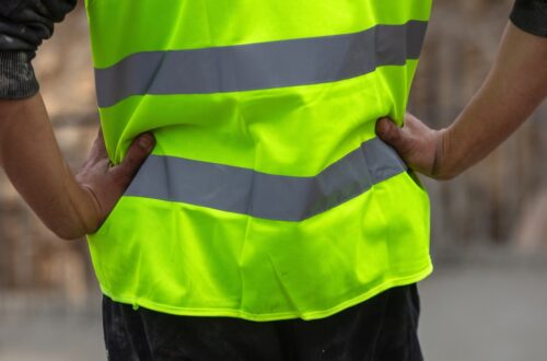 The view of someone from behind, with their hands on their hips, wearing a yellow high-visibility uniform during the day.