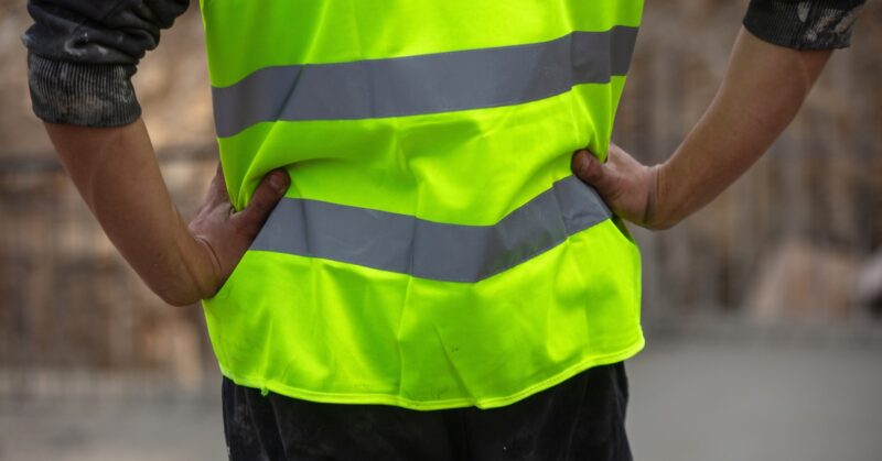 The view of someone from behind, with their hands on their hips, wearing a yellow high-visibility uniform during the day.