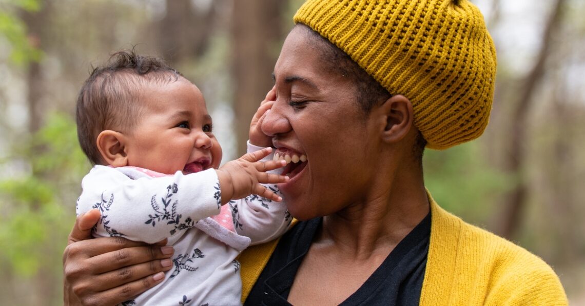 A woman dressed in a black shirt, yellow jacket, and yellow knitted beanie holding a baby while standing outside.