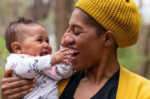A woman dressed in a black shirt, yellow jacket, and yellow knitted beanie holding a baby while standing outside.