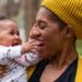 A woman dressed in a black shirt, yellow jacket, and yellow knitted beanie holding a baby while standing outside.