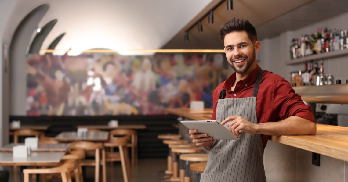 A restaurant owner in an apron holds a tablet at the bar, smiling in a modern dining room with wooden tables and stools.