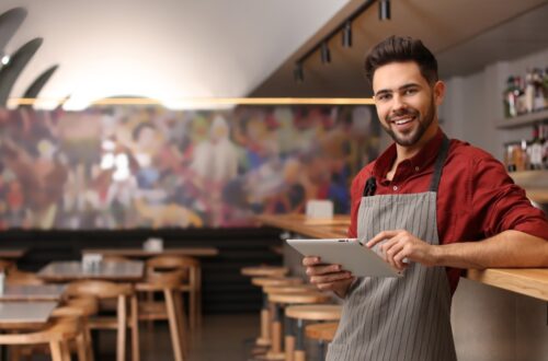 A restaurant owner in an apron holds a tablet at the bar, smiling in a modern dining room with wooden tables and stools.