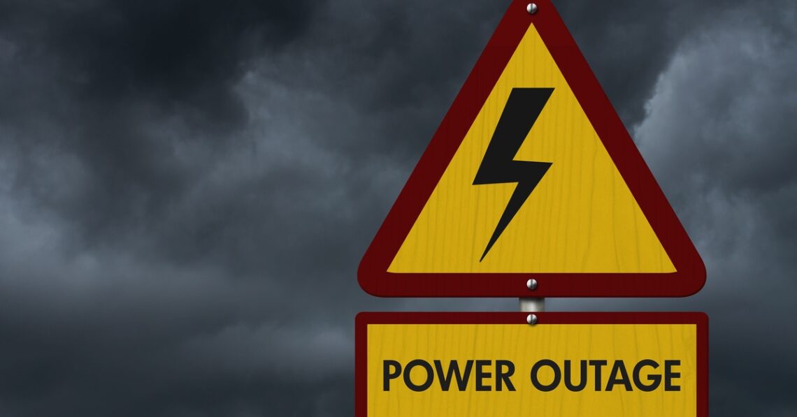 A yellow warning sign with a lightning bolt and “Power Outage” text against dark storm clouds in the background.