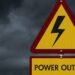 A yellow warning sign with a lightning bolt and “Power Outage” text against dark storm clouds in the background.