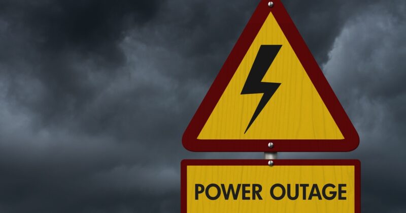 A yellow warning sign with a lightning bolt and “Power Outage” text against dark storm clouds in the background.