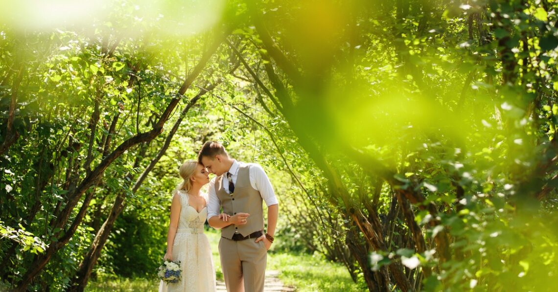 A bride in a white dress and a groom in a vest walk arm in arm along a leafy path in a bright green park.