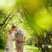 A bride in a white dress and a groom in a vest walk arm in arm along a leafy path in a bright green park.
