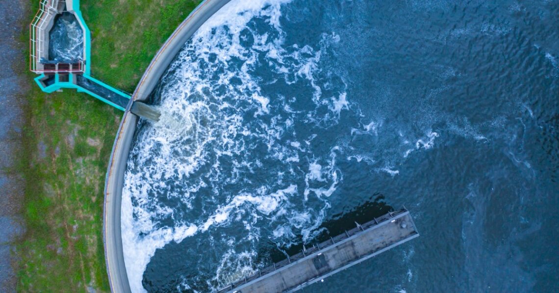 An aerial view of a full reservoir with blue, frothing water pouring into it. A long platform extends into the center.