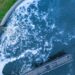 An aerial view of a full reservoir with blue, frothing water pouring into it. A long platform extends into the center.