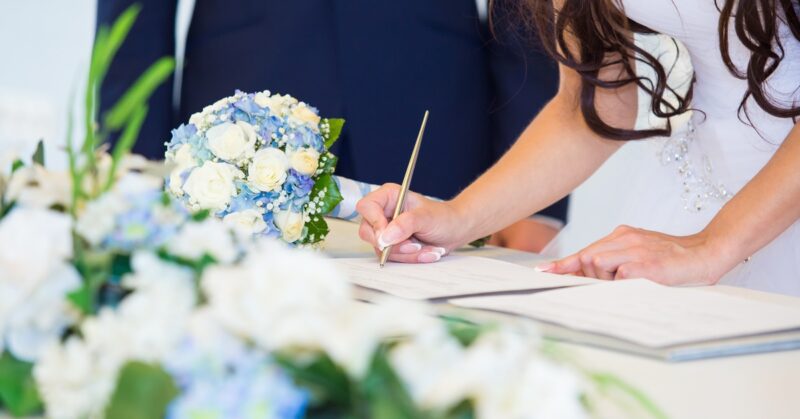 Bride signs a wedding license at a table, pen in hand, capturing a quiet, official moment after the ceremony.