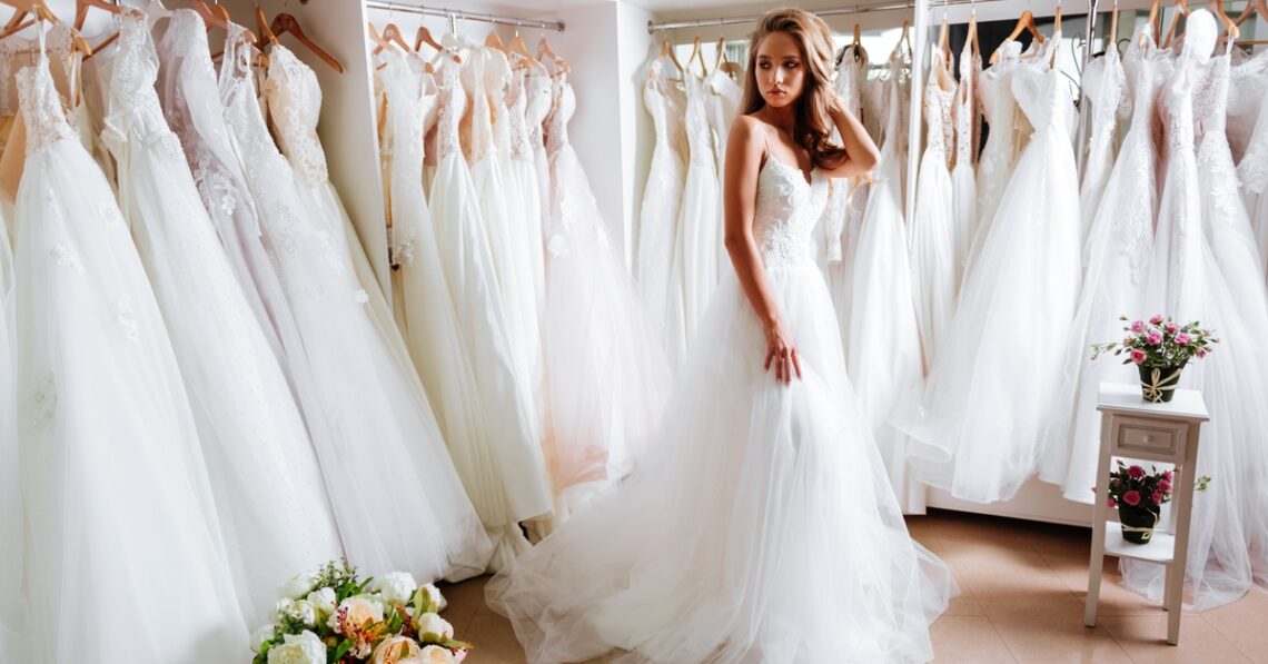 A young woman in a white wedding dress stands in a bridal boutique surrounded by rows of bridal gowns on display racks.