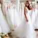 A young woman in a white wedding dress stands in a bridal boutique surrounded by rows of bridal gowns on display racks.