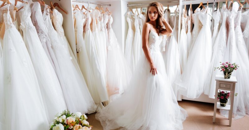 A young woman in a white wedding dress stands in a bridal boutique surrounded by rows of bridal gowns on display racks.