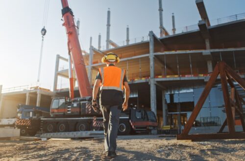 A person walking toward a jobsite while wearing an orange safety vest, a yellow hard hat, and yellow earmuffs.