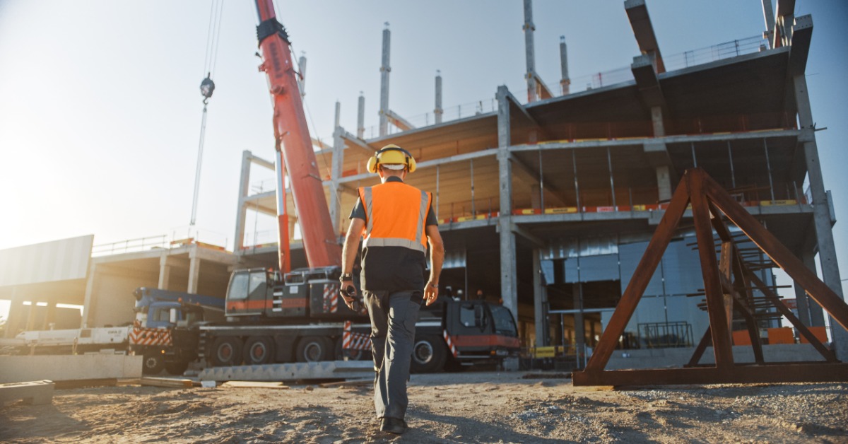 A person walking toward a jobsite while wearing an orange safety vest, a yellow hard hat, and yellow earmuffs.