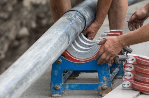 Two workers clamp a large gray pipe into a blue fusion machine with red jaws at a roadside trench.