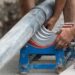 Two workers clamp a large gray pipe into a blue fusion machine with red jaws at a roadside trench.