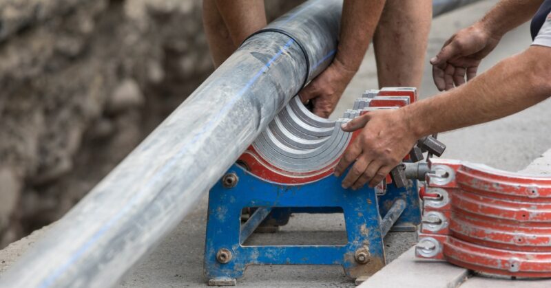 Two workers clamp a large gray pipe into a blue fusion machine with red jaws at a roadside trench.