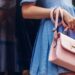 A woman in a blue dress holding a pink leather handbag with a gold clasp, leaning against a reflective wall.