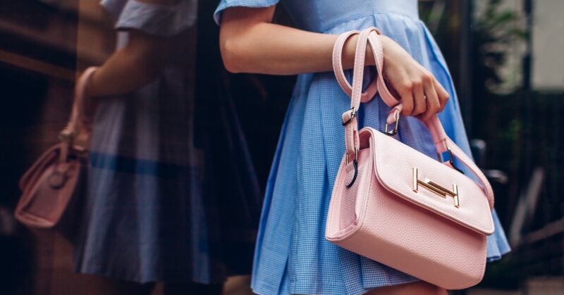 A woman in a blue dress holding a pink leather handbag with a gold clasp, leaning against a reflective wall.