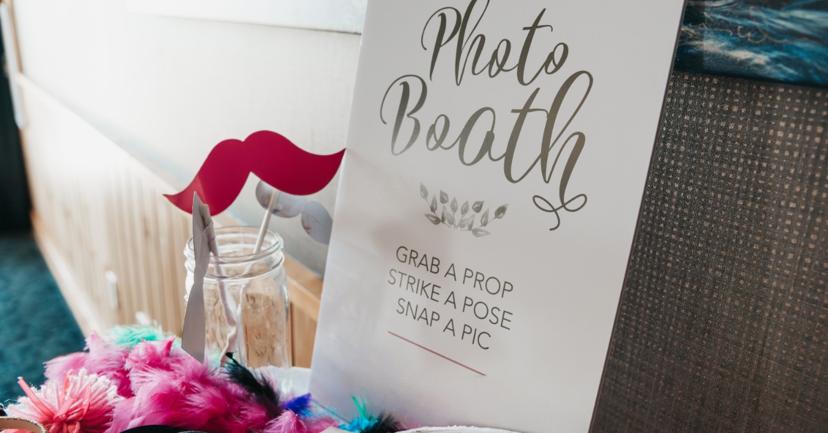 A photo booth setup with props beside a decorative sign reading “Photo Booth. Grab a prop, strike a pose, snap a pic”.