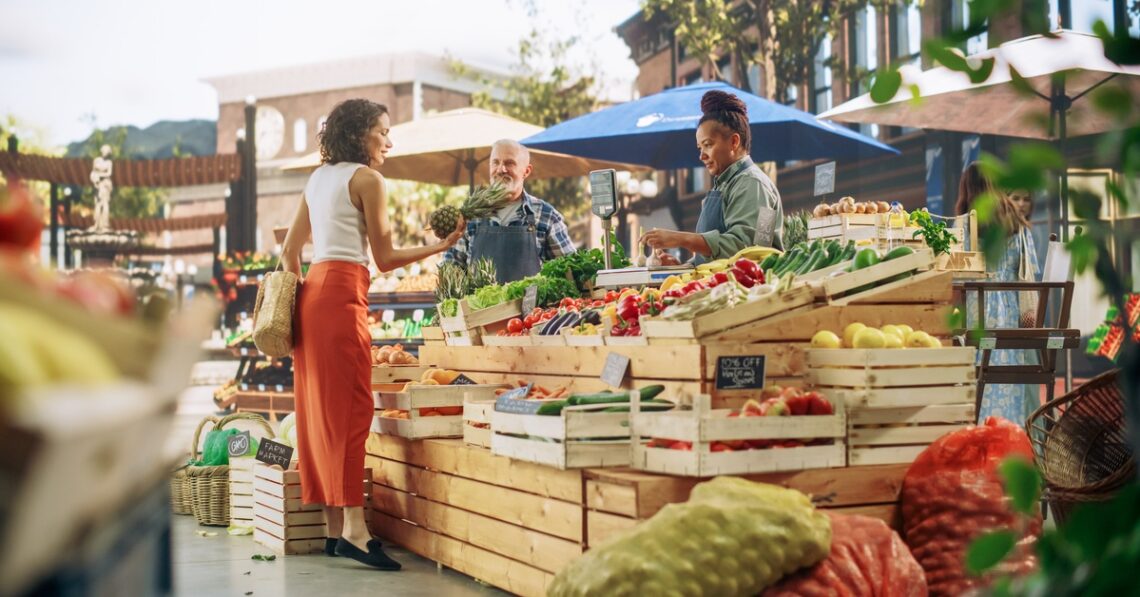 A woman stands at a farmers' market booth shopping for fresh produce. The booth is made out of wooden crates.