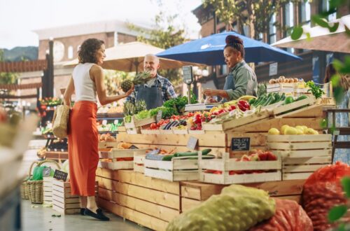 A woman stands at a farmers' market booth shopping for fresh produce. The booth is made out of wooden crates.