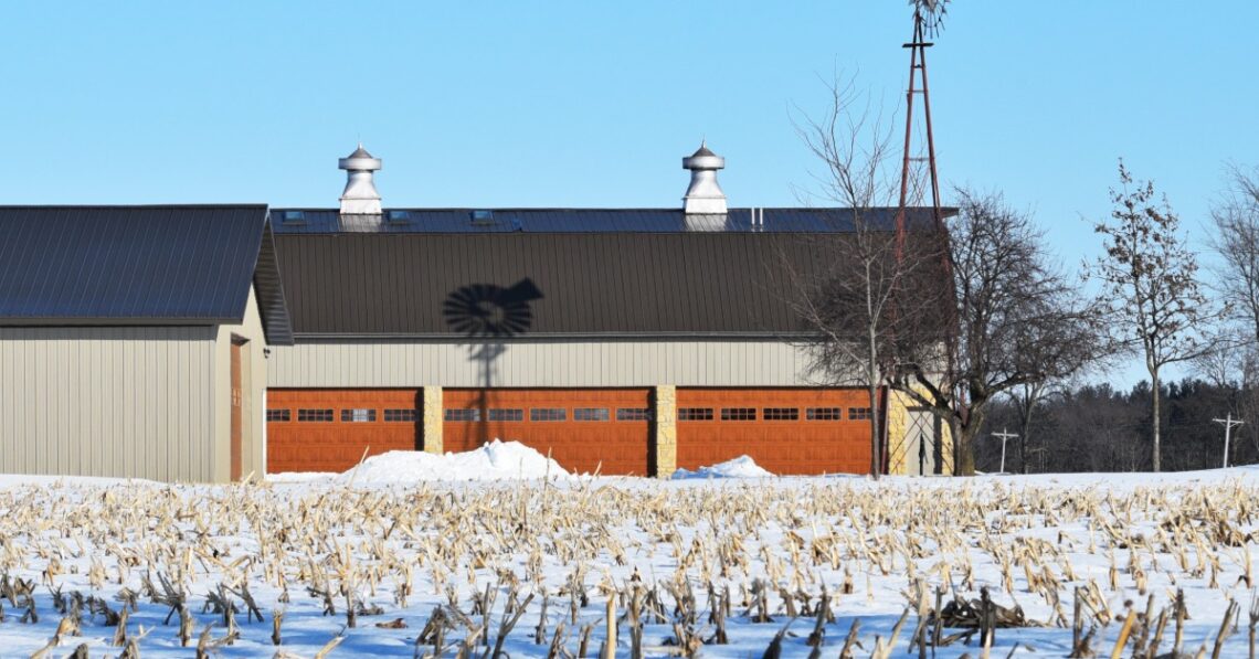 A windmill casts a shadow on a gray pole barn with red garage doors in a snowy field surrounded by trees.