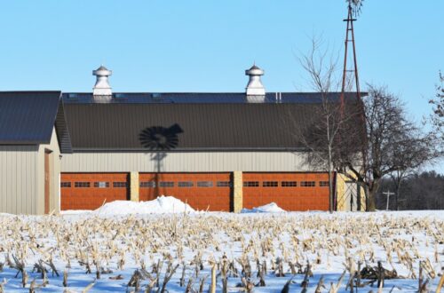 A windmill casts a shadow on a gray pole barn with red garage doors in a snowy field surrounded by trees.