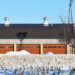 A windmill casts a shadow on a gray pole barn with red garage doors in a snowy field surrounded by trees.