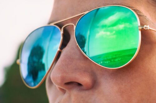 A close-up shows a person wearing reflective sunglasses that mirror a landscape and sky in blue and green tones.