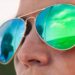 A close-up shows a person wearing reflective sunglasses that mirror a landscape and sky in blue and green tones.