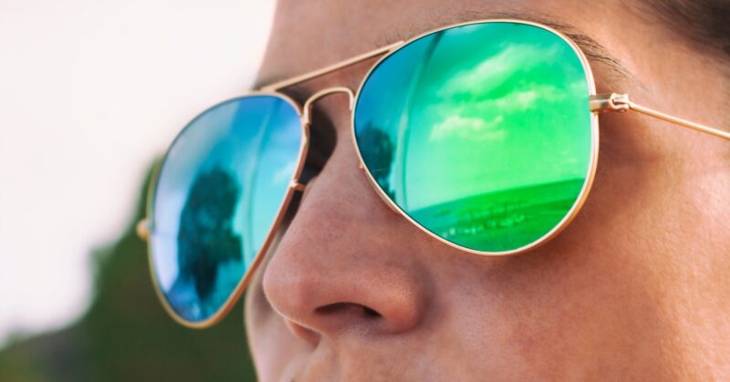 A close-up shows a person wearing reflective sunglasses that mirror a landscape and sky in blue and green tones.